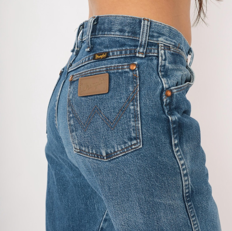Puede incluir: Vaqueros de mezclilla azules con un dise&ntilde;o cl&aacute;sico de cinco bolsillos. Los vaqueros presentan un parche de cuero marr&oacute;n con el logotipo "Wrangler" en el bolsillo trasero. Los vaqueros tienen remaches de cobre y cierre de bot&oacute;n.