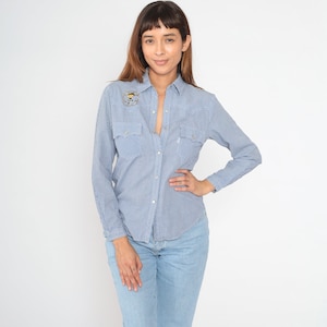 May include: A blue and white gingham button-down shirt with long sleeves and a small embroidered design on the left chest. The shirt has a pointed collar and two flap pockets on the chest.