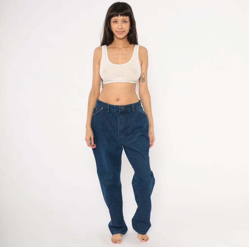 May include: A person wearing a white tank top and dark blue denim jeans. The jeans have a high waist and a relaxed fit. The person is standing in front of a white background.