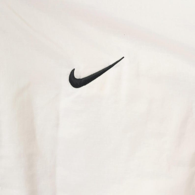 Puede incluir: Tejido blanco con un logotipo de Nike negro bordado en la esquina superior derecha. El tejido tiene un aspecto ligeramente texturizado.