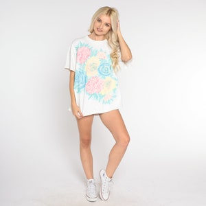 Puede incluir: Camiseta blanca de gran tama&ntilde;o con estampado floral en tonos rosa, azul y amarillo. Las flores son estilizadas y se asemejan a un ramo.