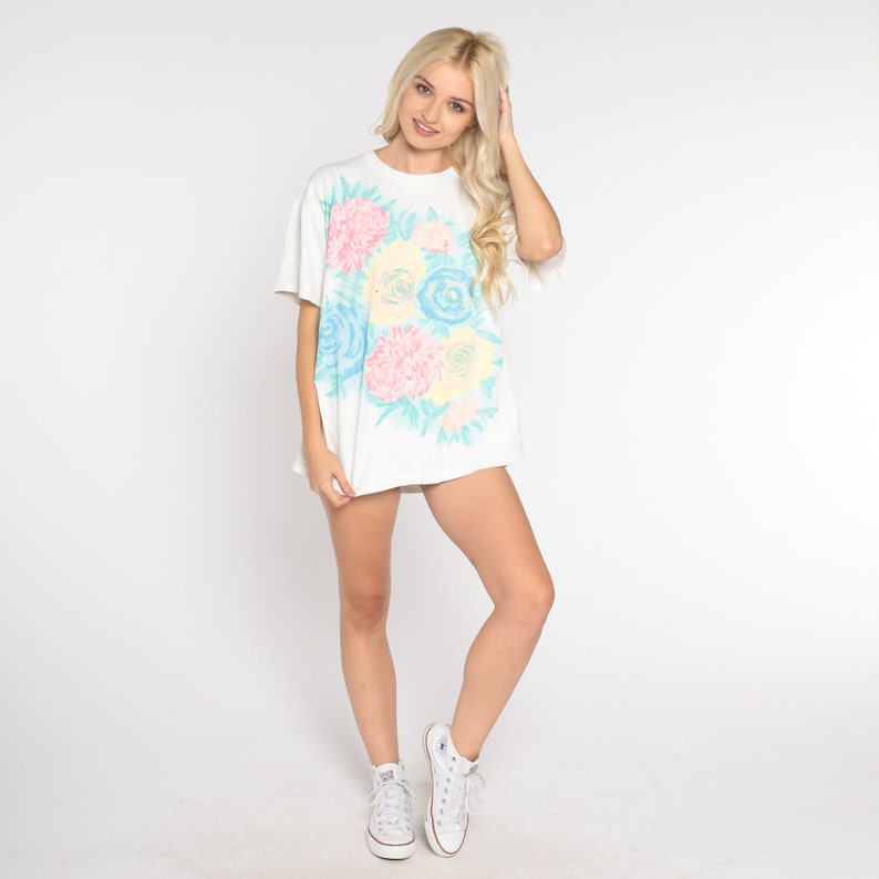 Puede incluir: Camiseta blanca de gran tama&ntilde;o con estampado floral en tonos rosa, azul y amarillo. Las flores son estilizadas y se asemejan a un ramo.