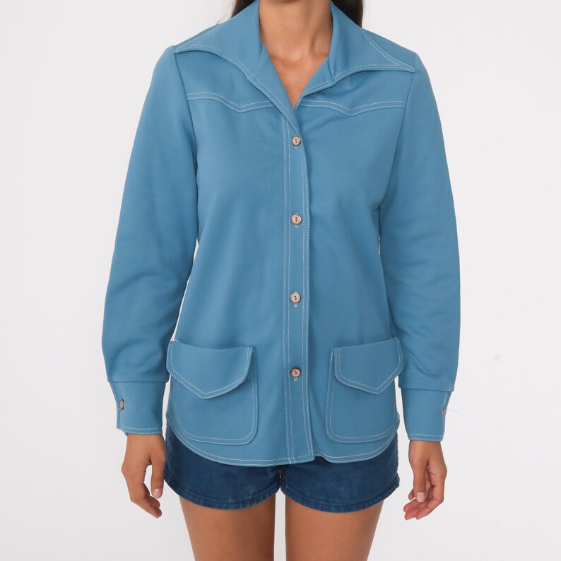 May include: A light blue long-sleeved shirt with a collared neckline, button closure, and two patch pockets. The shirt has white stitching details.