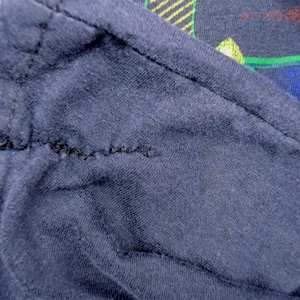 May include: Close-up of a navy blue fabric with a visible seam. The fabric appears to be a soft knit material. The background shows a glimpse of a colorful pattern with green, red, and blue elements.