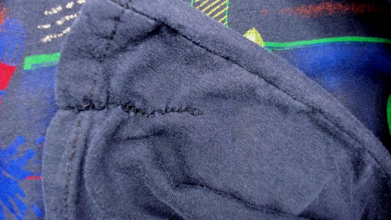 May include: Close-up of a navy blue fabric with a visible seam. The fabric appears to be a soft knit material. The background shows a glimpse of a colorful pattern with green, red, and blue elements.
