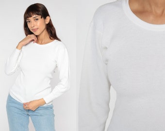 May include: A long-sleeve white thermal shirt featuring a crew neckline and a textured waffle-knit fabric pattern. The garment has fitted sleeves with ribbed cuffs and a straight hemline, providing a clean and simple look for everyday wear.