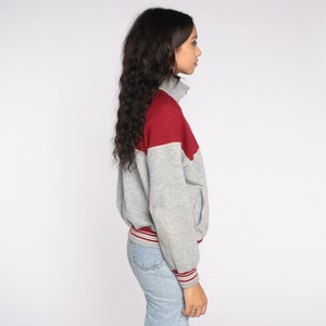 Op de afbeelding: Een grijze en bordeauxrode sweatshirt met ritssluiting. De sweatshirt heeft een bordeauxrode bovenborst en schoudergedeelte, met een witte bies. De mouwen en de body zijn grijs. De manchetten en de tailleband hebben bordeauxrode en witte strepen.