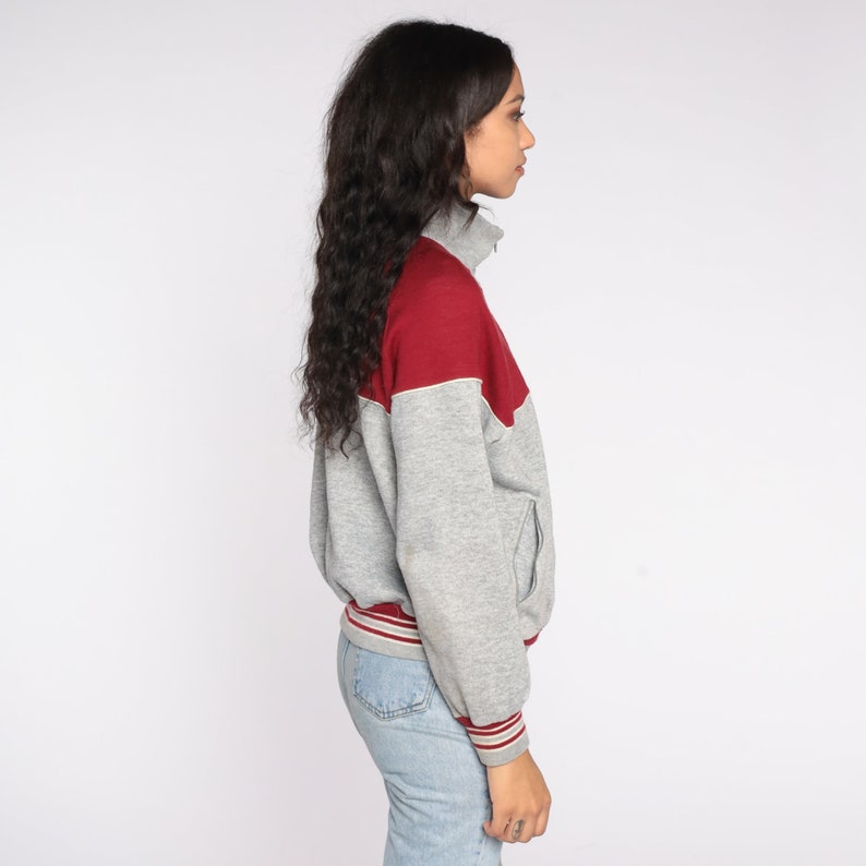 Op de afbeelding: Een grijze en bordeauxrode sweatshirt met ritssluiting. De sweatshirt heeft een bordeauxrode bovenborst en schoudergedeelte, met een witte bies. De mouwen en de body zijn grijs. De manchetten en de tailleband hebben bordeauxrode en witte strepen.