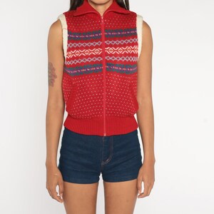 May include: Red sleeveless sweater vest with a white faux fur collar and a zip closure. The vest has a geometric pattern in blue, green, and white.