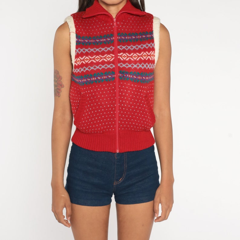 May include: Red sleeveless sweater vest with a white faux fur collar and a zip closure. The vest has a geometric pattern in blue, green, and white.