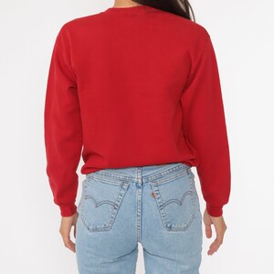 May include: A red long-sleeved sweatshirt with a crew neck. The sweatshirt is worn over a pair of light blue jeans.