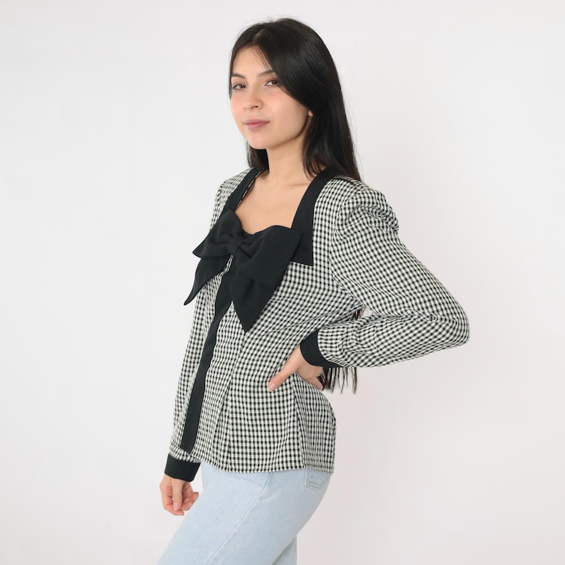 May include: A black and white gingham blouse with a large black bow at the neckline. The blouse has long sleeves and a fitted silhouette.