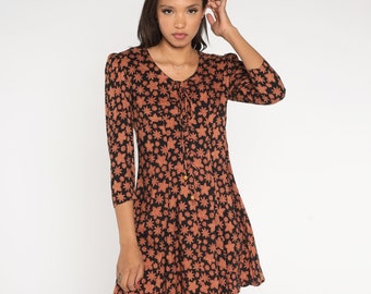Könnte beinhalten: Kurzes Kleid mit floralem Muster. Das Kleid hat einen schwarzen Hintergrund mit einem sich wiederholenden Muster aus orange-braunen Blumen. Es hat einen Rundhalsausschnitt mit Bindeband und einen ausgestellten Rock, mit Ärmeln, die bis zu den Ellbogen reichen.