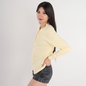 Puede incluir: Una camisa de manga larga de color amarillo pálido con cuello abotonado y un ajuste holgado. La camisa está hecha de una tela suave y cómoda y tiene un estilo casual.