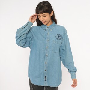 May include: Light blue denim button-down shirt with long sleeves. Features a small pocket on the chest and a navy blue patch with text and an emblem. The shirt has a classic collar and buttoned cuffs.
