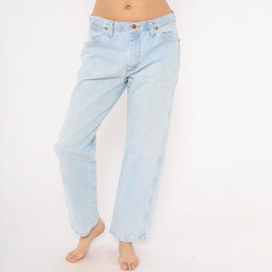 May include: Light blue denim jeans with a classic five-pocket design. The jeans have a straight leg cut and a faded wash, giving them a vintage look. The jeans are worn by a person.
