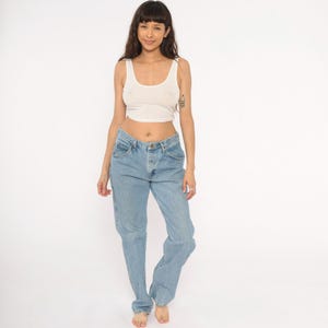 May include: A light-colored, sleeveless crop top and light blue denim jeans. The jeans have a classic five-pocket design and a relaxed fit. The model is standing in front of a white background.