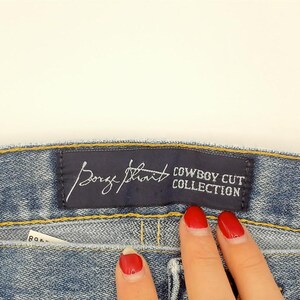Peut inclure: Gros plan sur un jean en denim bleu avec une &eacute;tiquette bleu fonc&eacute; indiquant "Borge Strait COWBOY CUT COLLECTION". Le jean a un design classique avec des coutures visibles et un d&eacute;lavage clair.