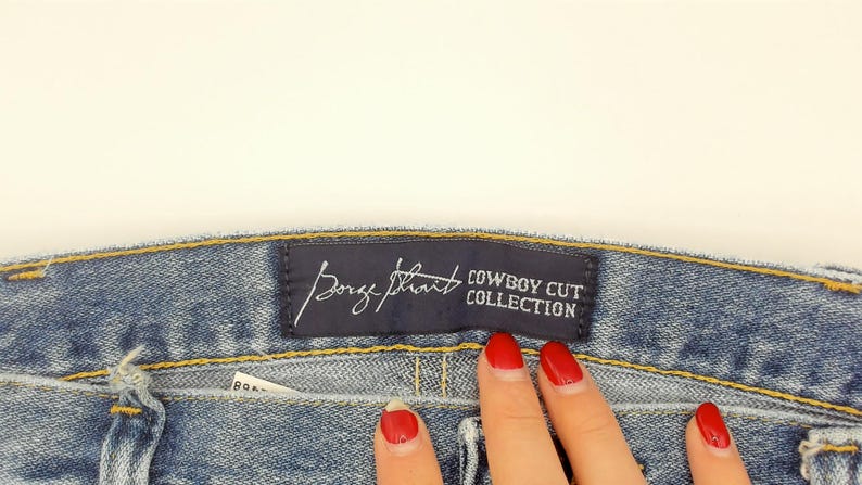 Peut inclure: Gros plan sur un jean en denim bleu avec une &eacute;tiquette bleu fonc&eacute; indiquant "Borge Strait COWBOY CUT COLLECTION". Le jean a un design classique avec des coutures visibles et un d&eacute;lavage clair.