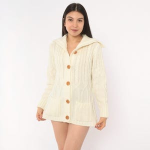 May include: A cream-colored cable knit cardigan featuring a wide collar, long sleeves, and a front button closure with five round brown buttons. The garment includes two patch pockets on the lower front and a textured vertical cable knit pattern.