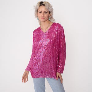 Pode incluir: Uma blusa magenta de lantejoulas de manga comprida com decote em V. A blusa é coberta por pequenas lantejoulas brilhantes, criando um efeito texturizado e brilhante. A peça é usada com jeans azul claro.