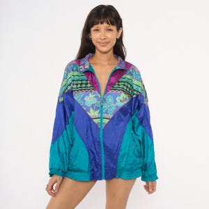 May include: A long-sleeve zip-up windbreaker jacket featuring a vibrant color-blocked design in shades of royal blue, teal, and purple. The upper chest and collar area display a multi-colored floral and geometric pattern with green accents.