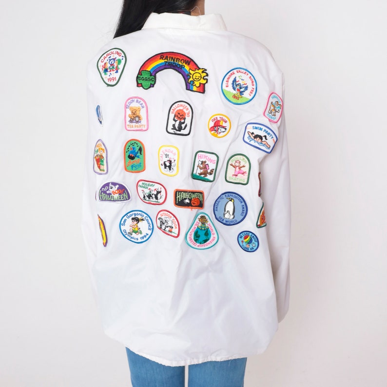 Op de afbeelding: Wit overhemd met lange mouwen en knopen met een verscheidenheid aan kleurrijke geborduurde patches. De patches hebben verschillende ontwerpen, waaronder dieren, feestdagen en activiteiten. De patches zijn op de achterkant van het overhemd genaaid.