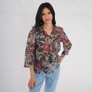 May include: A woman wearing a long-sleeved, button-up shirt with a floral print in shades of red, brown, and black. The shirt has a relaxed fit and a collar.