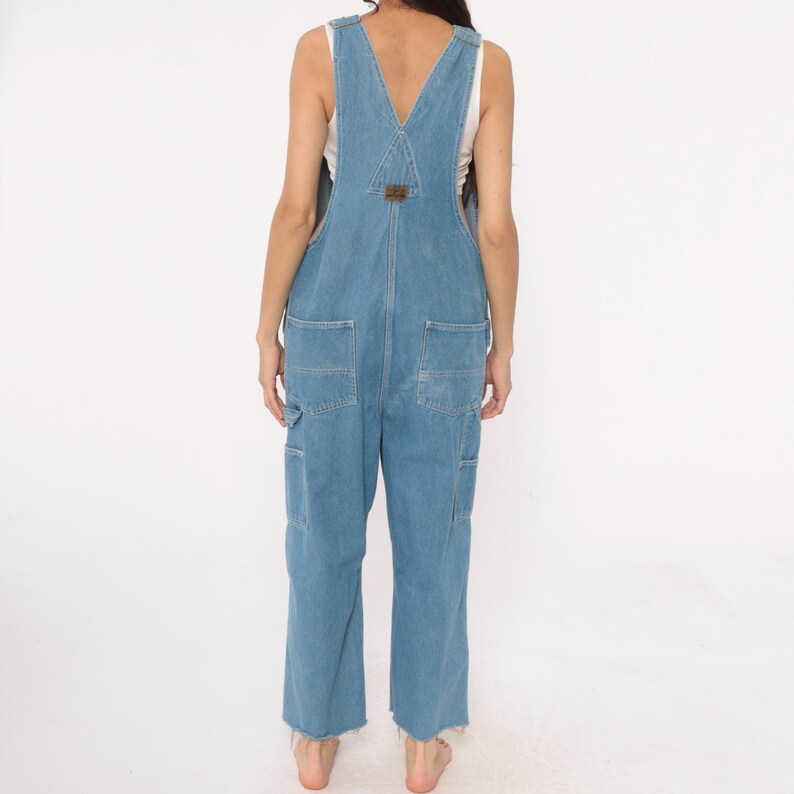 May include: Light blue denim overalls with a V-neck and multiple pockets. The overalls have a relaxed fit and a raw hem. A small brown patch is sewn on the bib. The model is wearing a white tank top underneath.