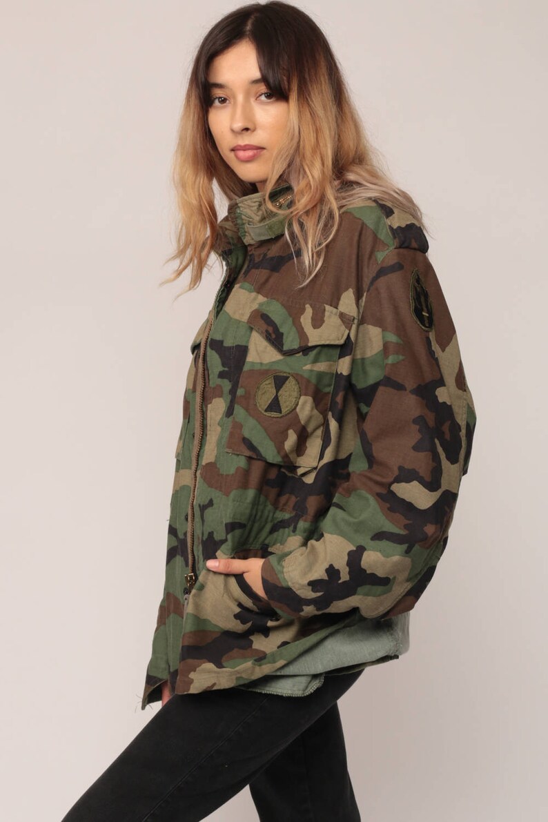 May include: A camouflage field jacket featuring a classic woodland pattern in shades of brown, green, and black. The jacket includes a high collar, a front zipper closure, two chest pockets with flaps, and circular patches on the chest and sleeve.