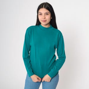 Pode incluir: Camisa de manga comprida verde-azulado com gola alta. A camisa &eacute; feita de um material macio. A modelo est&aacute; usando jeans azuis. A camisa &eacute; de cor s&oacute;lida e tem um design simples.