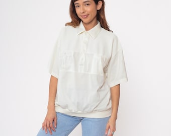 Puede incluir: Camisa crema de manga corta con cuello y dos bolsillos en el pecho. La camisa tiene una tapeta con botones y cintura fruncida. La modelo lleva vaqueros azul claro.