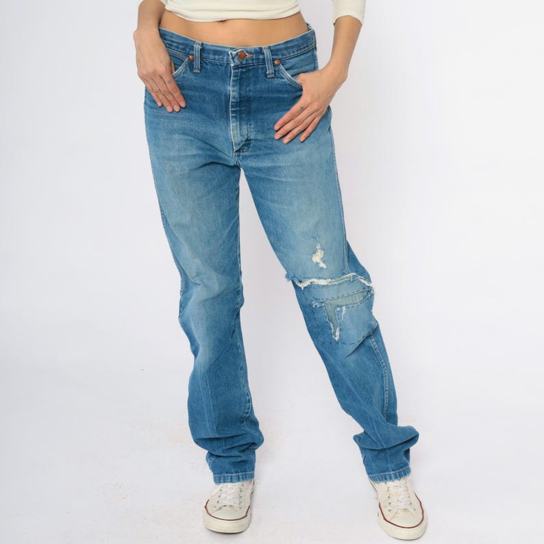 May include: Light blue denim jeans with distressed detailing, including a ripped knee. The jeans have a classic five-pocket design and a straight leg fit. The model is wearing white trainers.