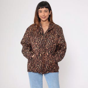 Puede incluir: Una chaqueta de estampado animal marrón y beige con cierre de cremallera. La chaqueta tiene dos bolsillos en la parte delantera.