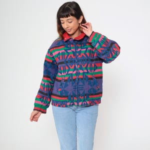 May include: A vintage, zip-up jacket with a geometric pattern in shades of blue, pink, green, and black. The jacket has a red collar and cuffs. The model is wearing light blue jeans.