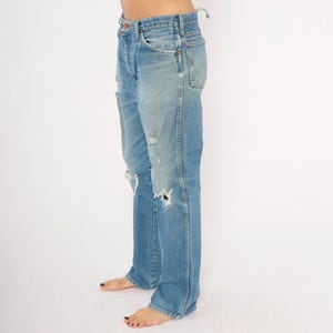May include: Light blue denim jeans with distressed details, including rips and faded patches. The jeans have a classic five-pocket design and a straight leg fit. The jeans are worn and have a vintage look.