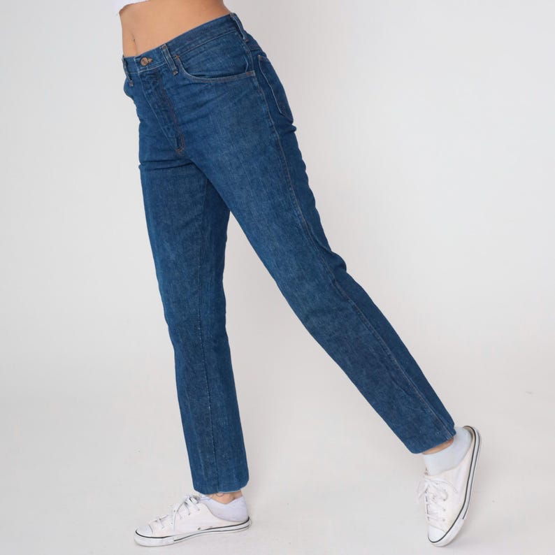 May include: Dark blue denim jeans with a classic straight leg fit. The jeans have a button and zipper closure, front and back pockets, and a faded wash. The jeans are paired with white sneakers.