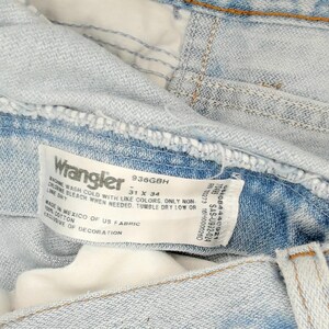 Op de afbeelding: Close-up van een lichtblauwe Wrangler-jeans. De jeans heeft een vervaagde wassing en zichtbare stiksels. Een wit label toont het Wrangler-logo, maat 79 x 86 cm en wasvoorschriften. Gemaakt in Mexico van Amerikaanse stof.