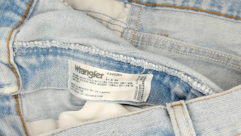 Op de afbeelding: Close-up van een lichtblauwe Wrangler-jeans. De jeans heeft een vervaagde wassing en zichtbare stiksels. Een wit label toont het Wrangler-logo, maat 79 x 86 cm en wasvoorschriften. Gemaakt in Mexico van Amerikaanse stof.