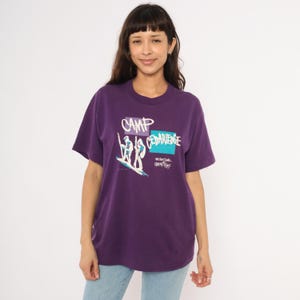 May include: A purple t-shirt with the words "CAMP CEDARIDGE" in white and blue. The shirt features a graphic of two figures on a surfboard. The shirt is a crew neck and has short sleeves.