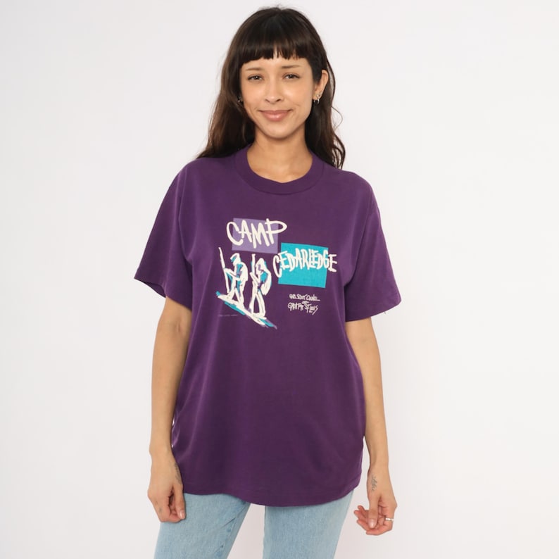 May include: A purple t-shirt with the words "CAMP CEDARIDGE" in white and blue. The shirt features a graphic of two figures on a surfboard. The shirt is a crew neck and has short sleeves.