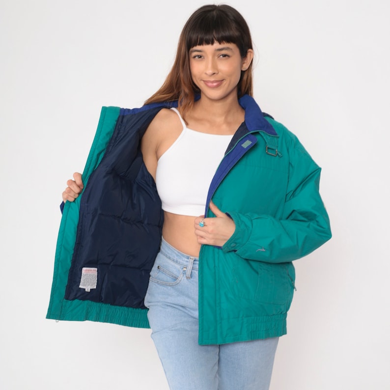 K&ouml;nnte beinhalten: Eine reversible Jacke in Tealblau und Marineblau mit Rei&szlig;verschluss und Druckknopfverschluss. Die Jacke hat eine Kapuze und zwei Taschen auf der Vorderseite.