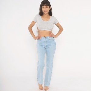 Op de afbeelding: Een vrouw draagt een lichtgrijze crop top met korte mouwen en lichtblauwe jeans. De jeans heeft een hoge taille en rechte pijpen. Ze staat met haar handen in haar zij tegen een witte achtergrond.
