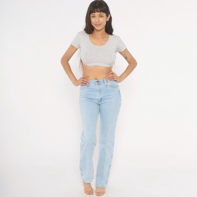 Op de afbeelding: Een vrouw draagt een lichtgrijze crop top met korte mouwen en lichtblauwe jeans. De jeans heeft een hoge taille en rechte pijpen. Ze staat met haar handen in haar zij tegen een witte achtergrond.
