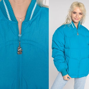 May include: A turquoise blue puffer jacket with a zipper closure and a silver swan charm. The jacket has a white and turquoise blue striped collar.