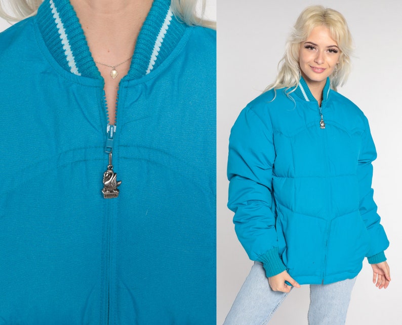 May include: A turquoise blue puffer jacket with a zipper closure and a silver swan charm. The jacket has a white and turquoise blue striped collar.