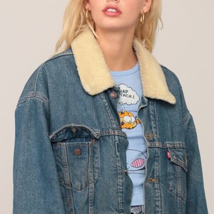 Puede incluir: Una chaqueta de mezclilla azul con cuello de piel sint&eacute;tica color crema, botones de metal y dos bolsillos en el pecho con solapa. Una peque&ntilde;a etiqueta roja es visible en el bolsillo izquierdo. La chaqueta se lleva sobre una camiseta azul claro.