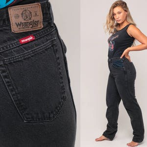 May include: Black Wrangler jeans with a classic fit. The jeans feature a brown leather patch with the Wrangler logo and a red logo tag on the back pocket. The model is wearing a black tank top.