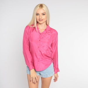 Könnte beinhalten: Eine Frau trägt ein rosa Satinhemd mit Knöpfen, langen Ärmeln und Jeans-Shorts.