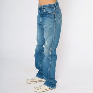 May include: Faded blue denim jeans with distressed patches on the knees. The jeans are worn with white canvas trainers. The overall style is casual and relaxed.